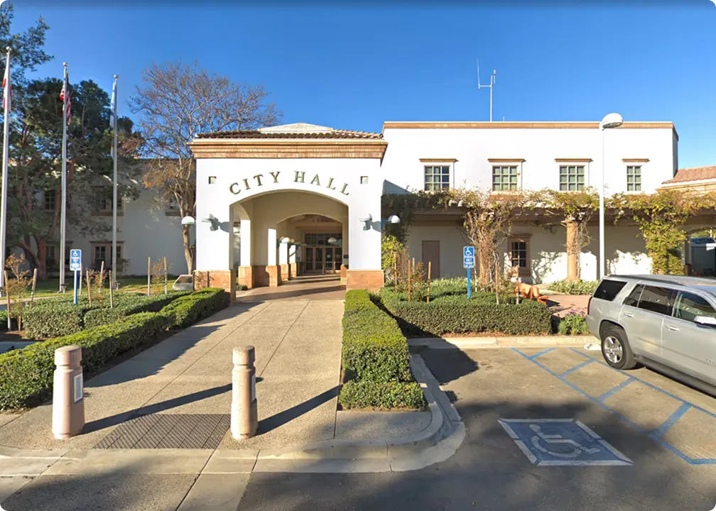 White city hall building with an arched entrance and landscaped hedges along the pathway, with a clear blue sky above.