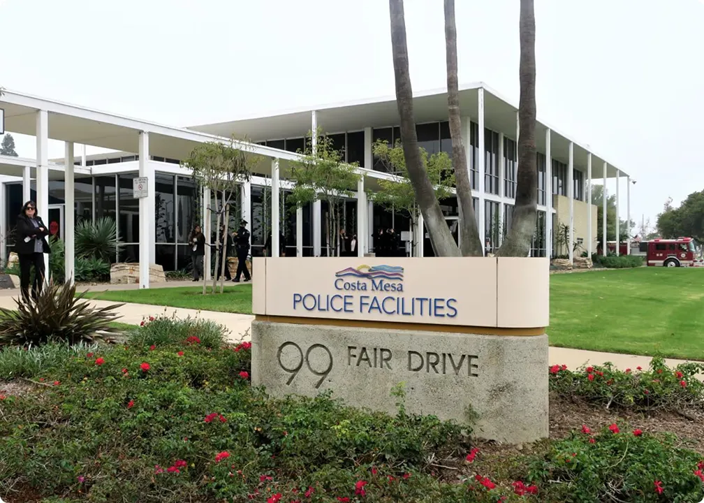 Exterior view of Costa Mesa Police Facilities building with a sign showing the address 99 Fair Drive surrounded by landscaping and palm trees.