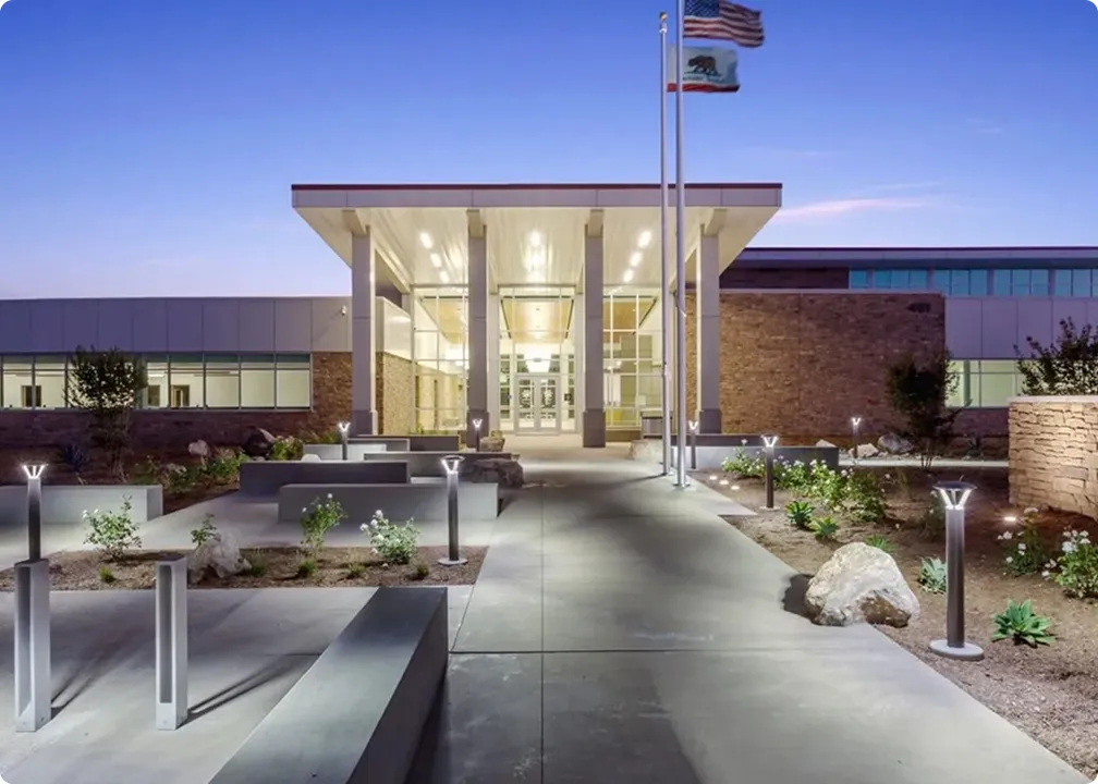 Modern building entrance with tall columns, illuminated pathway lights, and American and California state flags at dusk.