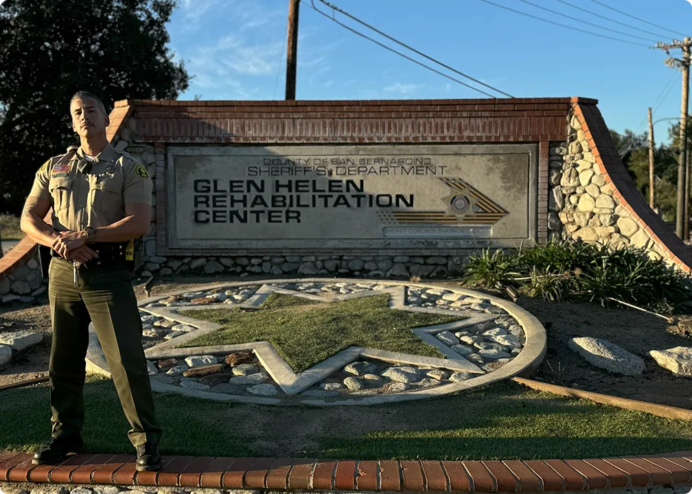 Uniformed sheriff officer standing with arms crossed in front of Glen Helen Rehabilitation Center stone sign and landscaped star emblem.