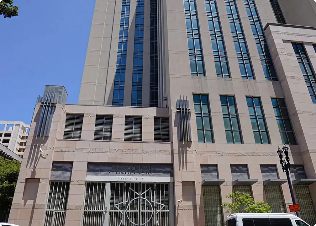Tall beige building with large vertical windows under a clear blue sky, with security bars and a white vehicle partially visible in front.