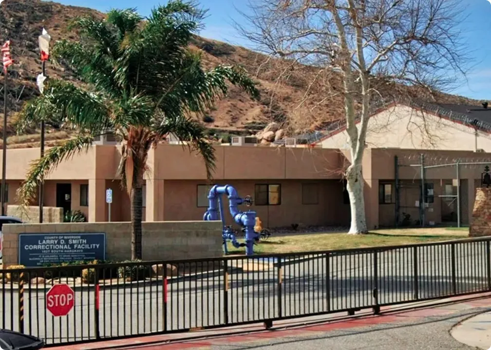 Exterior view of Larry D. Smith Correctional Facility with a palm tree, a leafless tree, and a black metal fence in front under a clear sky.