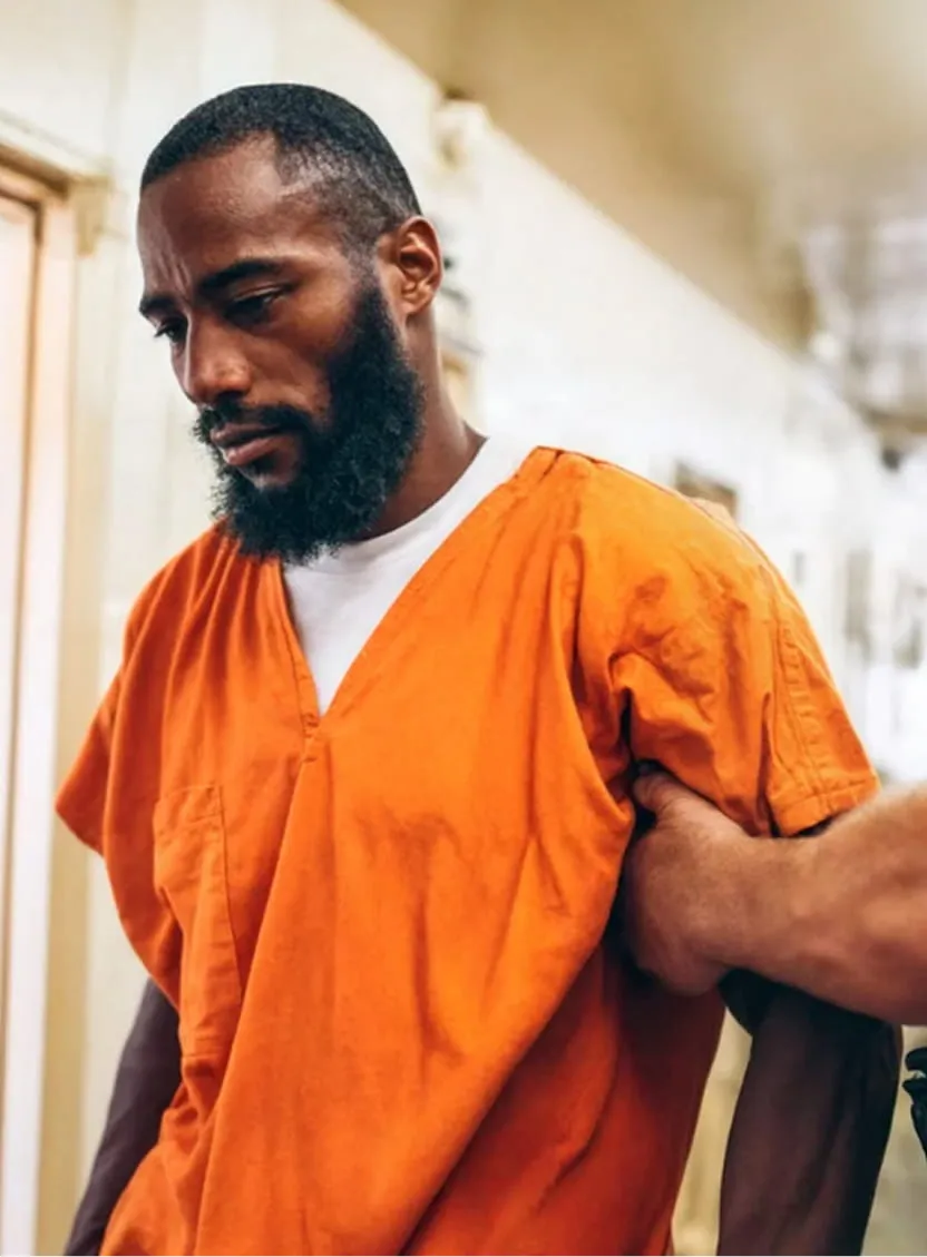 Man in orange prison uniform being escorted by a guard holding his arm in a corridor.
