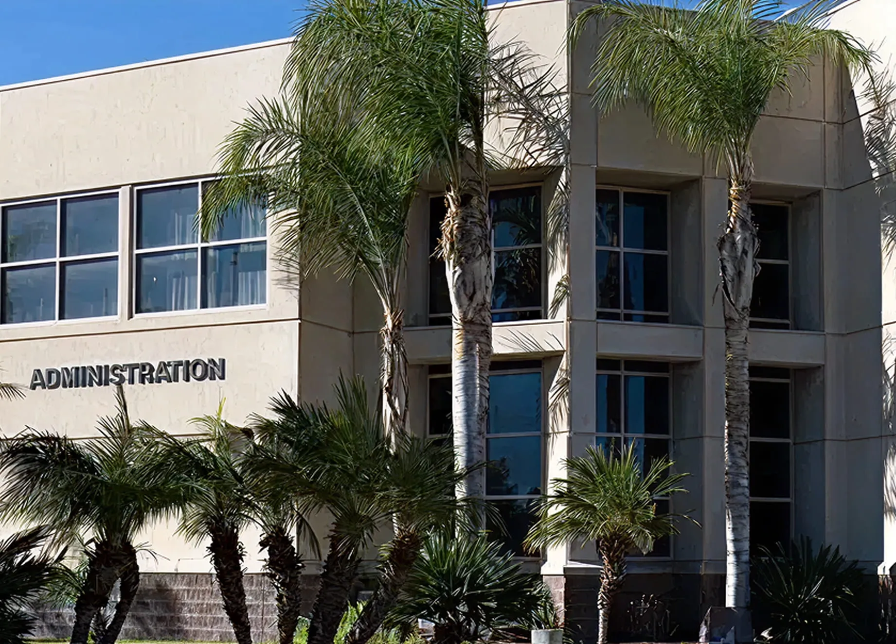 Exterior view of a beige administration building with large windows and several palm trees in front.