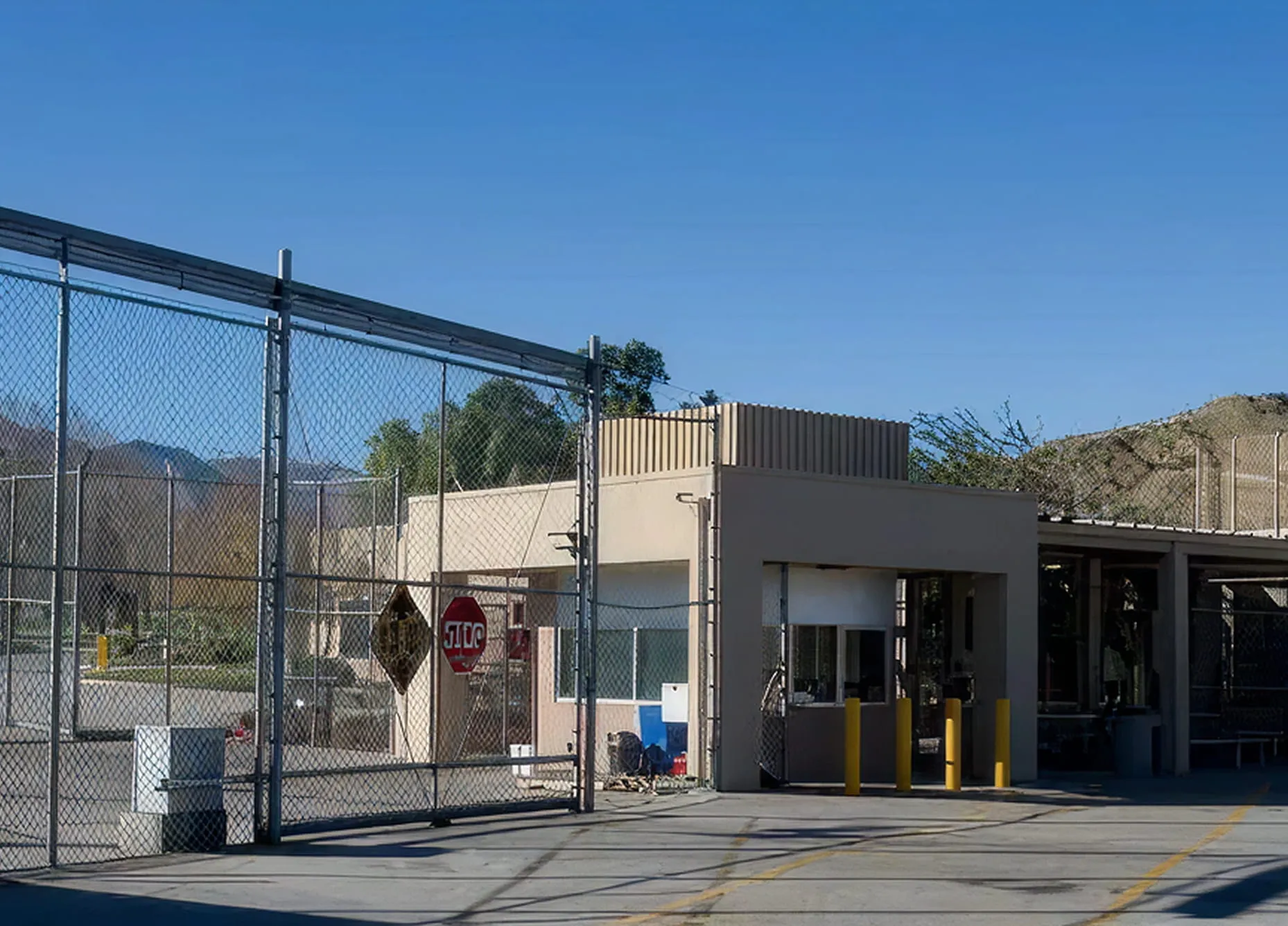 Security checkpoint with a beige guardhouse, chain-link fences, and a stop sign under a clear blue sky.