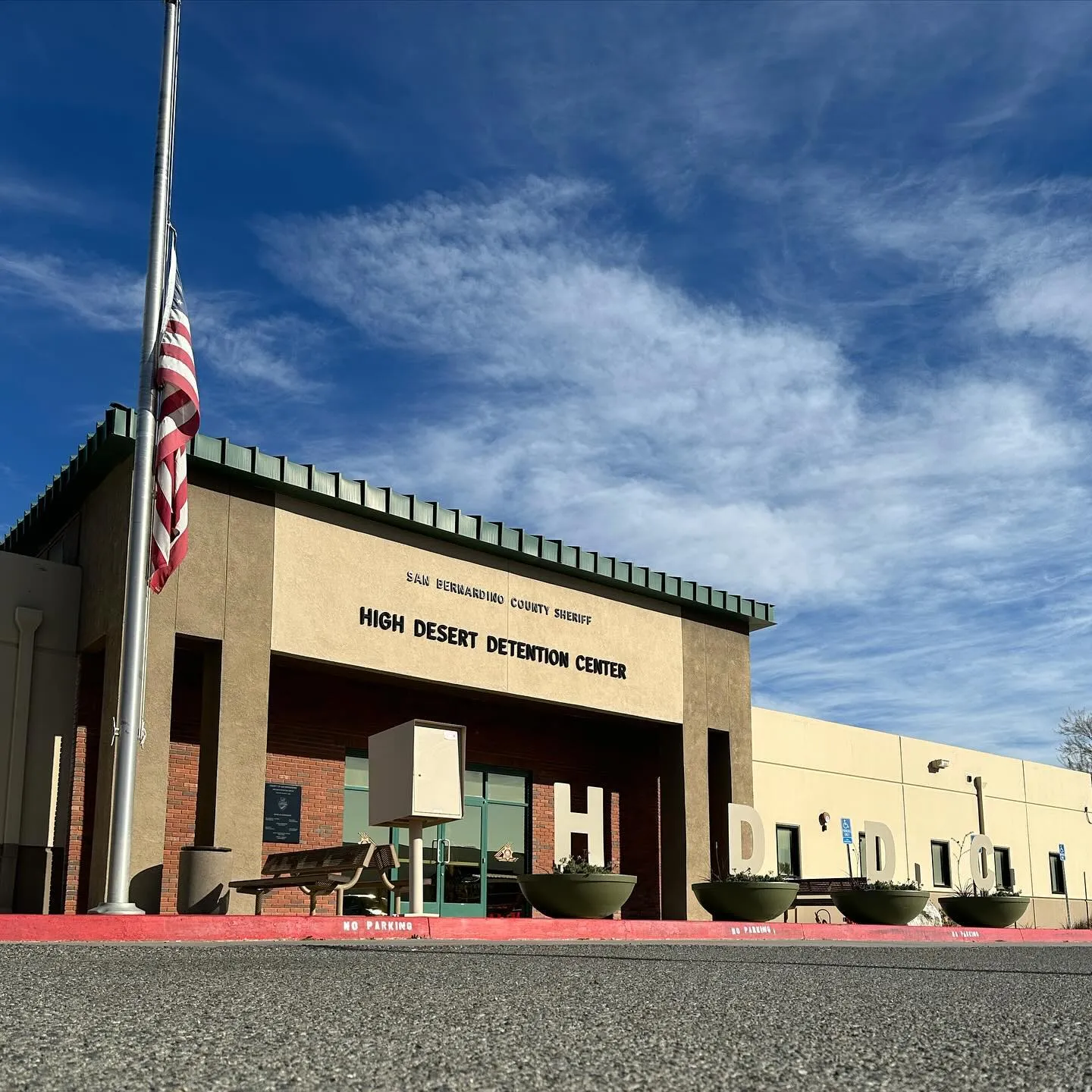 Front entrance of the High Desert Detention Center with an American flag at half-mast and large letters spelling H D D C in planters.