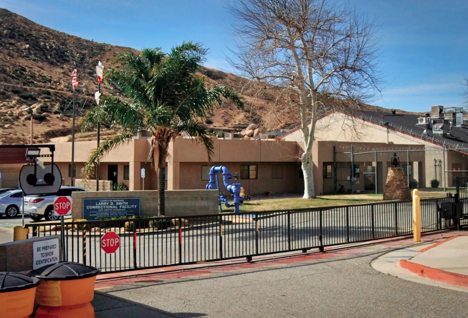 Entrance gate of Larry D. Smith Correctional Facility with stop signs, palm tree, and a building in a mountainous desert area.
