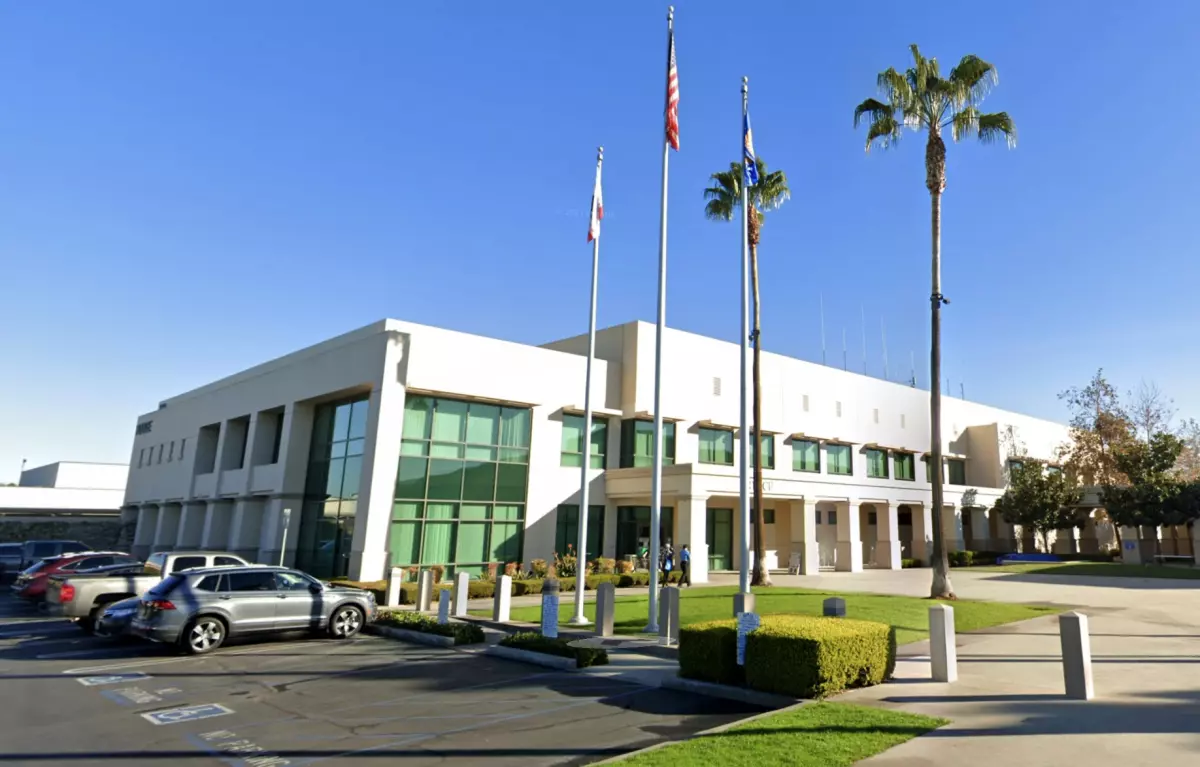 Modern two-story white government building with green-tinted windows, three flagpoles, palm trees, and parked cars in front.