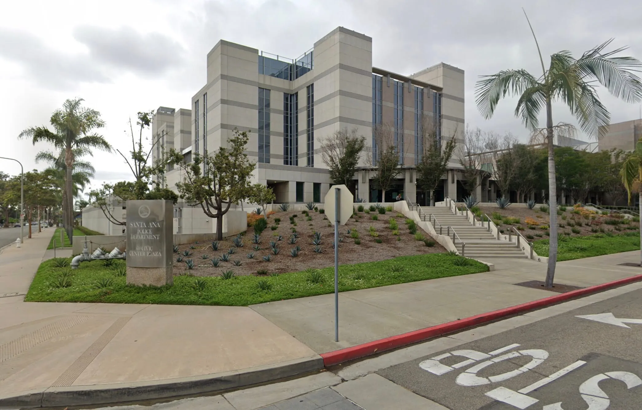 Modern multi-story building with large windows and a sign reading Santa Ana Police Department Civic Center Plaza at the front, surrounded by trees and landscaped greenery.
