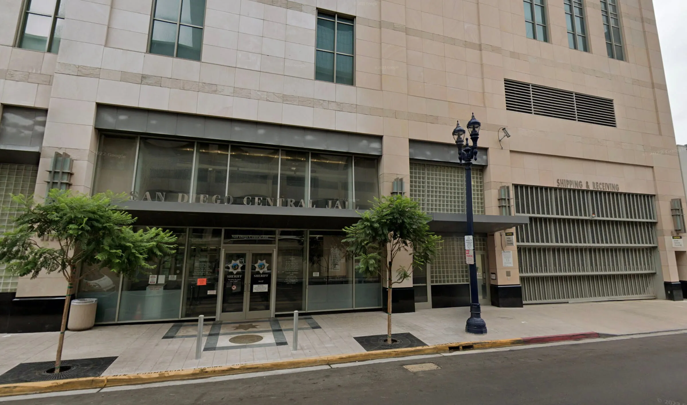 Exterior entrance of San Diego Central Jail with glass doors, two small trees, and a streetlamp.