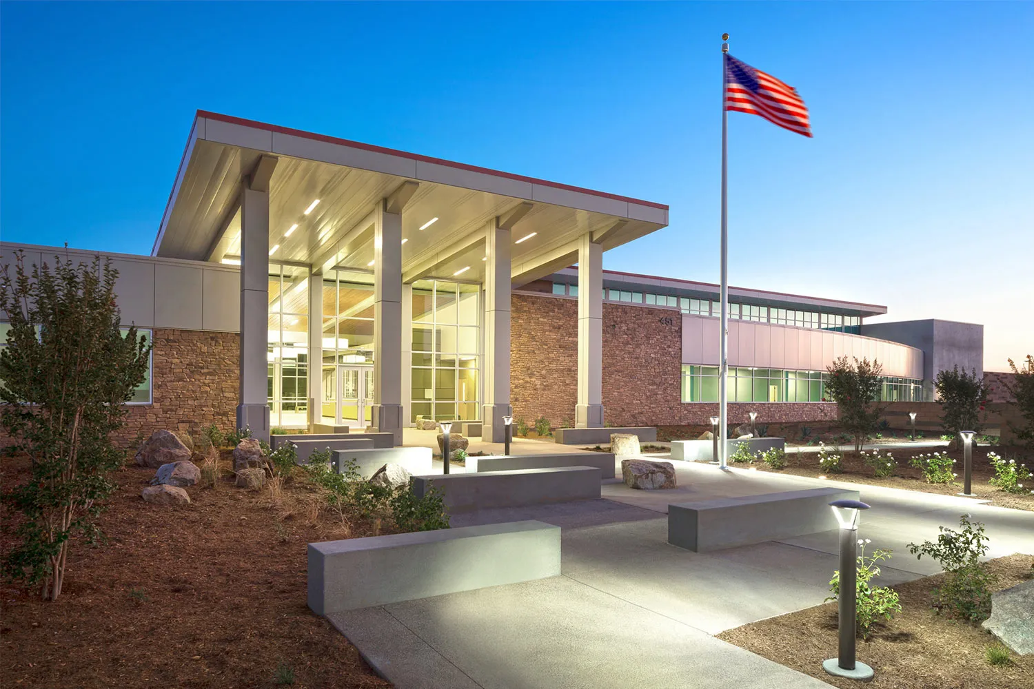 Modern building entrance with tall pillars, illuminated pathway, landscaping, and an American flag on a flagpole at dusk.
