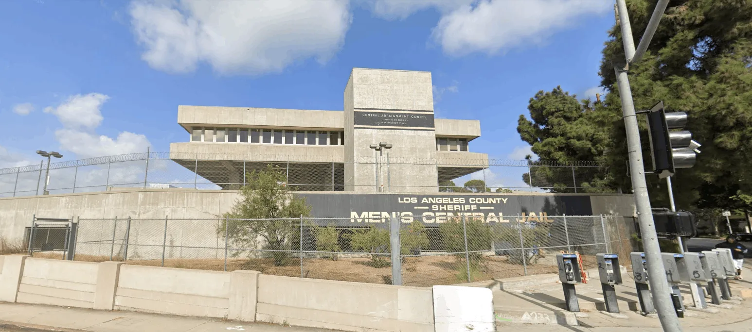Exterior view of Los Angeles County Sheriff's Men’s Central Jail with concrete building, barbed wire fence, and payphones on the sidewalk.