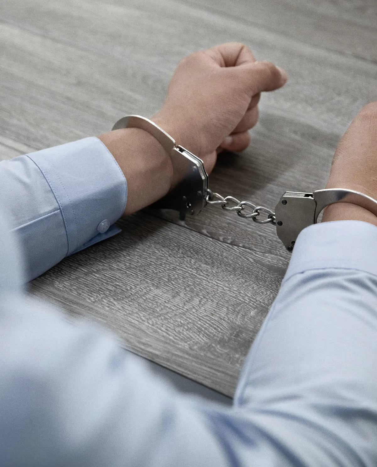 Person wearing a light blue shirt with hands cuffed together in metal handcuffs on a wooden surface.