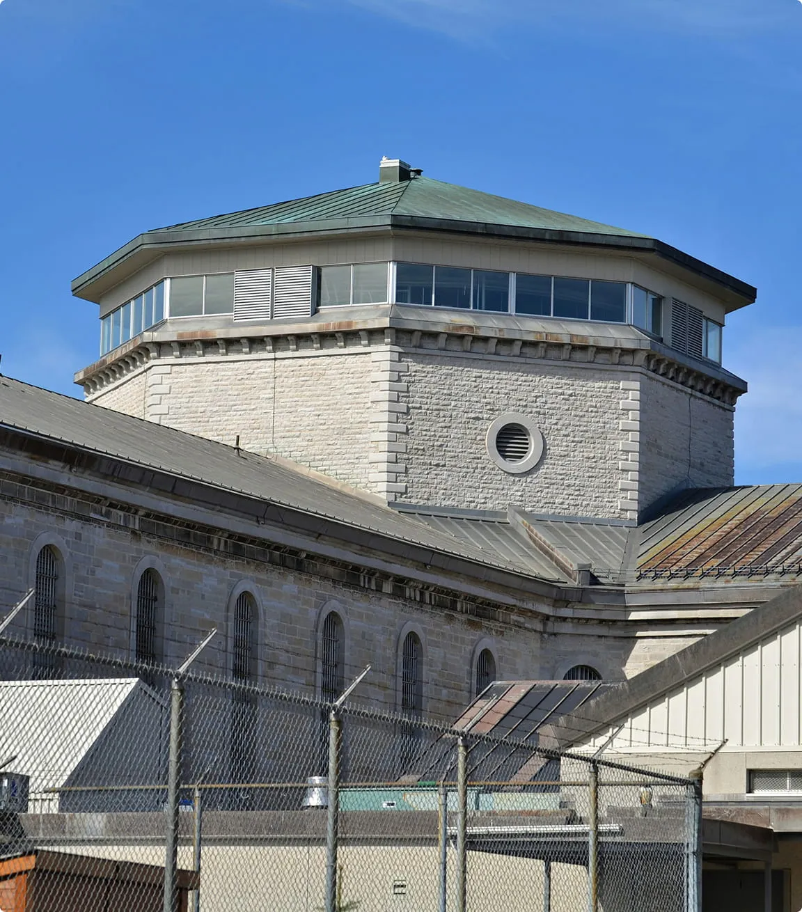 Stone prison building with barred windows, octagonal watchtower, and chain-link fence topped with barbed wire under a clear blue sky.