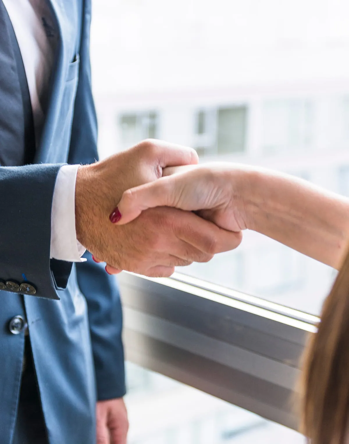 Close-up of a handshake between a man in a blue suit and a woman with painted nails near a window.