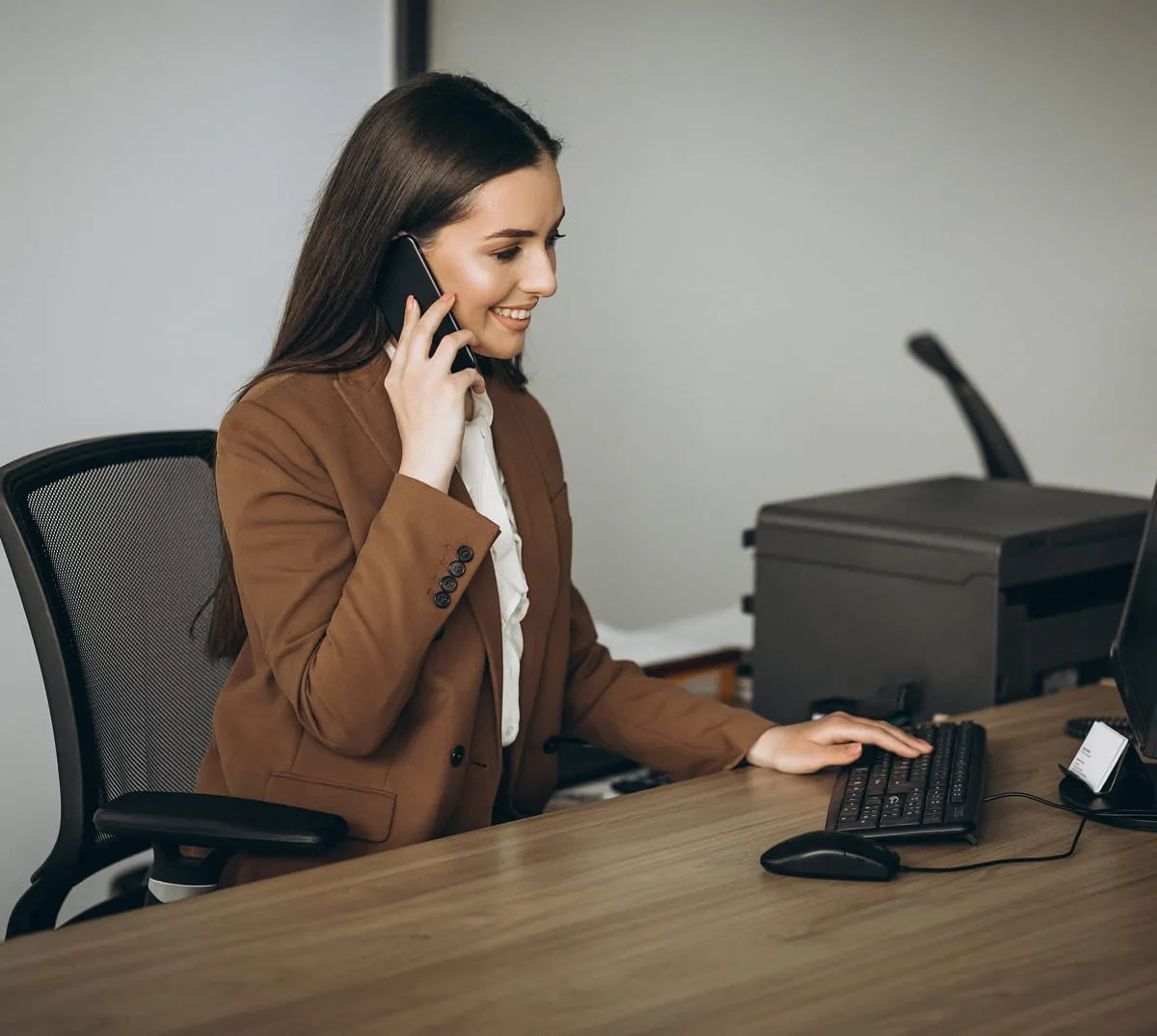 Smiling woman in brown blazer talking on the phone while typing on a computer keyboard at an office desk.