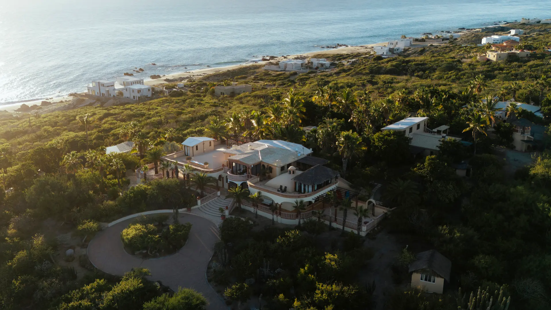 Aerial view of a large coastal house surrounded by dense greenery with the ocean and beach in the background.