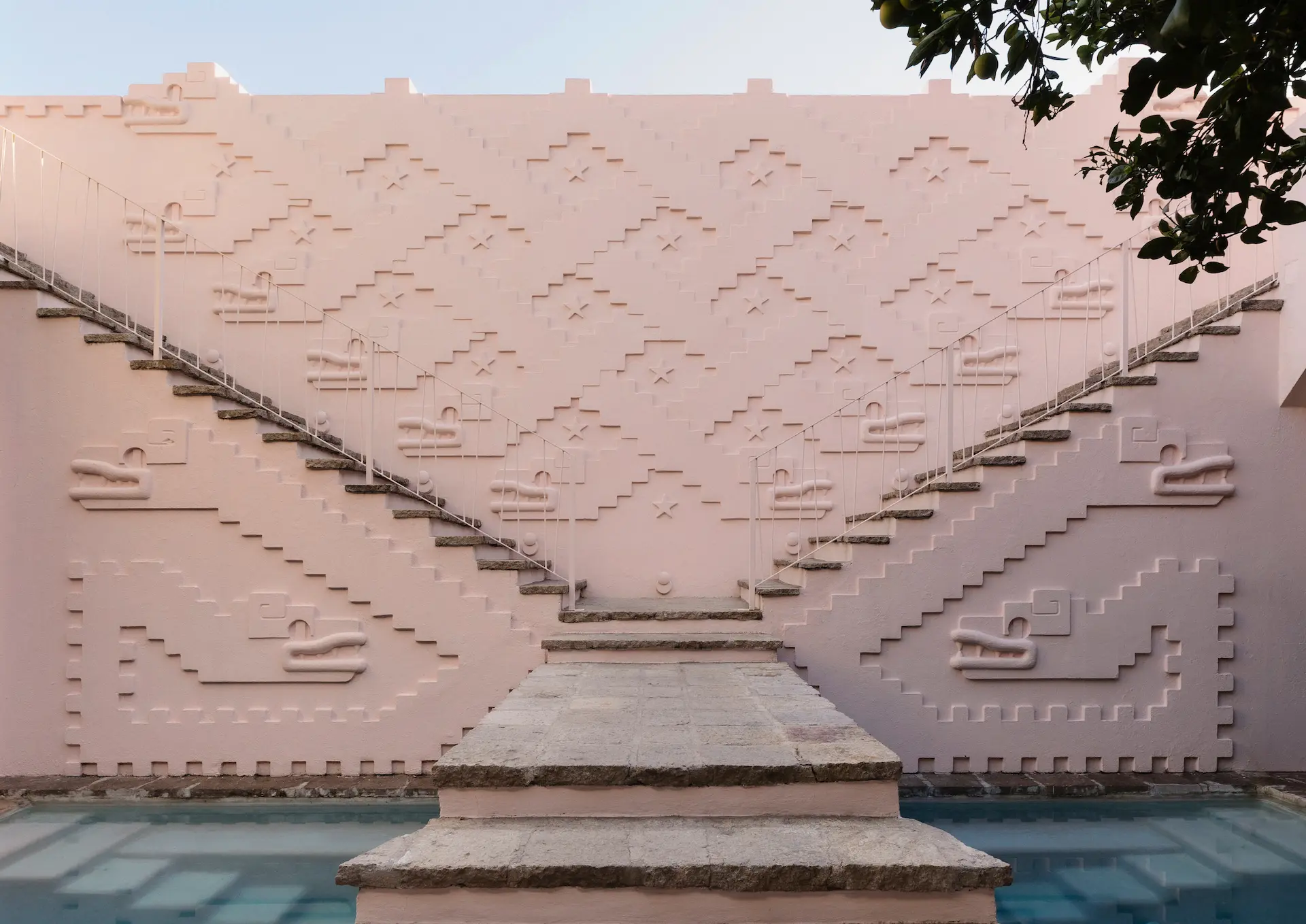 Symmetrical pink staircases with geometric and animal-like reliefs on the walls, leading up from a reflective pool.