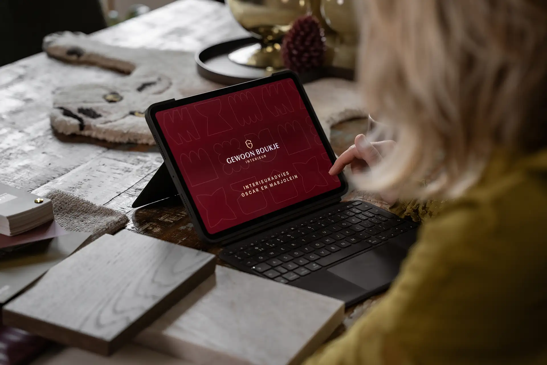 Persoon kijkt op een tablet met het logo van Gewoon Boukje Interieur en tekst over interieuradvies op een houten tafel met kleurstalen.