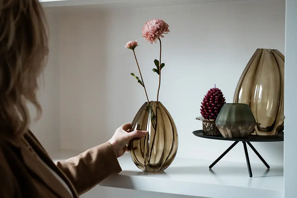 Hand plaatst een beige transparante vaas met twee roze bloemen op een witte plank naast andere vazen en decoraties.