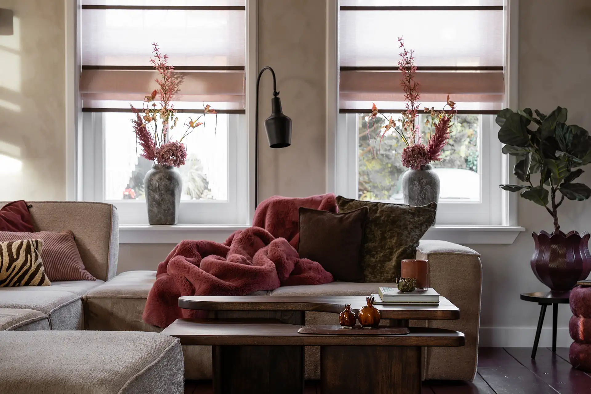 Gezellige woonkamer met een beige hoekbank, een roze gebreide deken, kussens, bloemen in vazen op de vensterbank en een houten salontafel met decoraties.