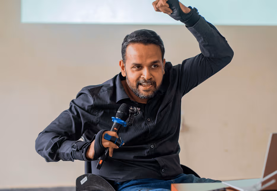 Man in a wheelchair giving an enthusiastic presentation, holding a microphone and raising one arm.