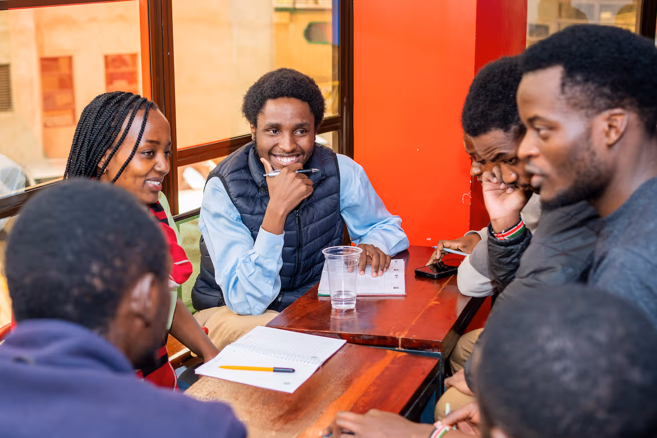 Group of young adults sitting around a table indoors, engaged in discussion with notebooks and a smartphone.