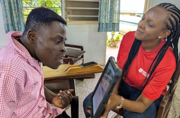 A woman in a red shirt showing a tablet to a man in a pink checkered shirt inside a room with chairs and curtains.