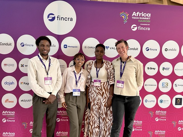 A group of four people standing in front of a bright pink event backdrop featuring numerous sponsor logos for the Africa Tech Summit Nairobi powered by Fincra. They are wearing conference badges and lanyards, and posing together for a group photo. The back