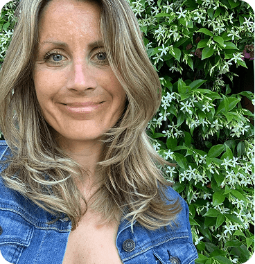 Vrouw met blond haar en blauwe spijkerjas die lacht, met een achtergrond van groen gebladerte en witte bloemen.