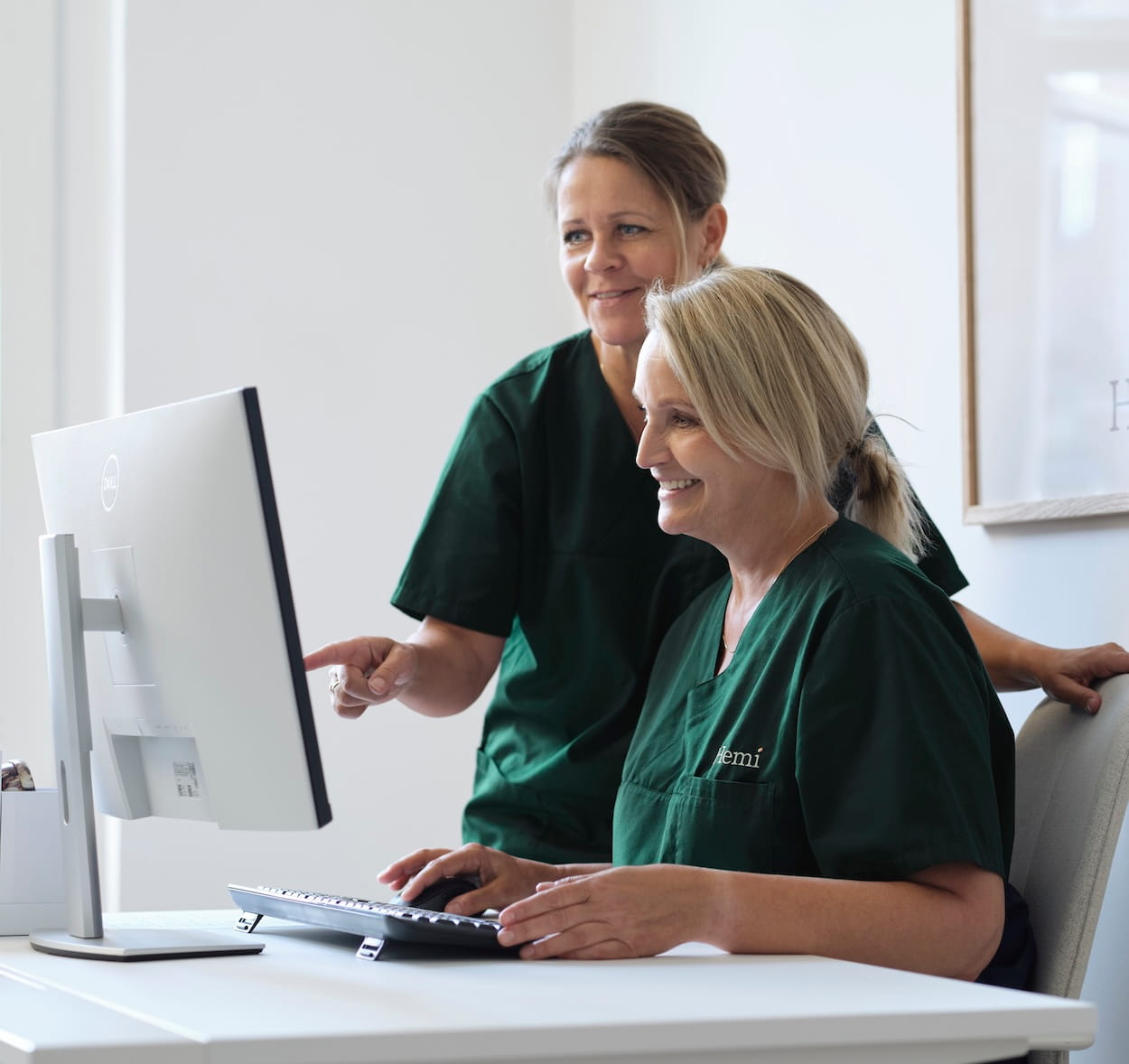 Twee vrouwen in groene medische kleding werken samen aan een computerscherm in een lichte kantooromgeving.