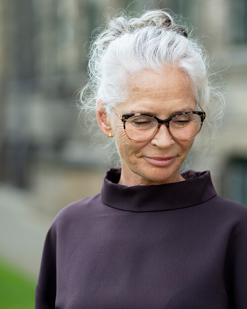 Oudere vrouw met grijs haar in een nonchalante knot, glazen en een donkerkleurige coltrui, kijkt glimlachend omlaag buiten.
