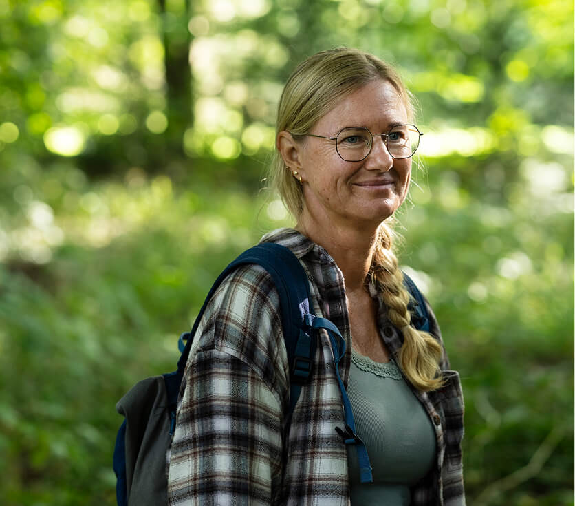 Vrouw met bril en rugzak, lachend in een bosrijke omgeving met groene bladeren op de achtergrond.