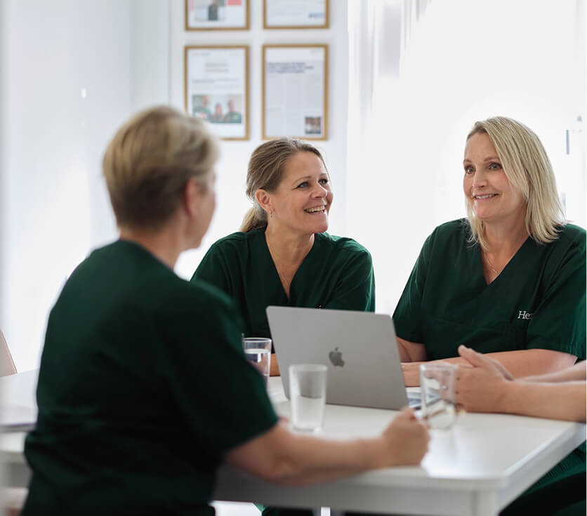 Drie vrouwen in groene uniformen zitten samen aan een tafel met een laptop en glazen water, in een lichte kamer.