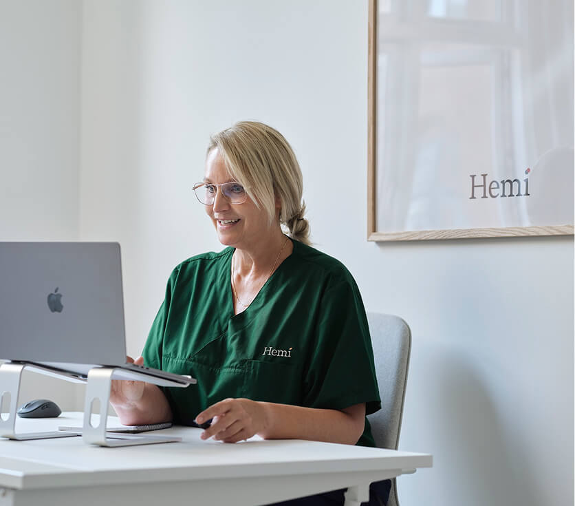 Vrouw in groene medische outfit die glimlacht tijdens het werken op een laptop aan een wit bureau.