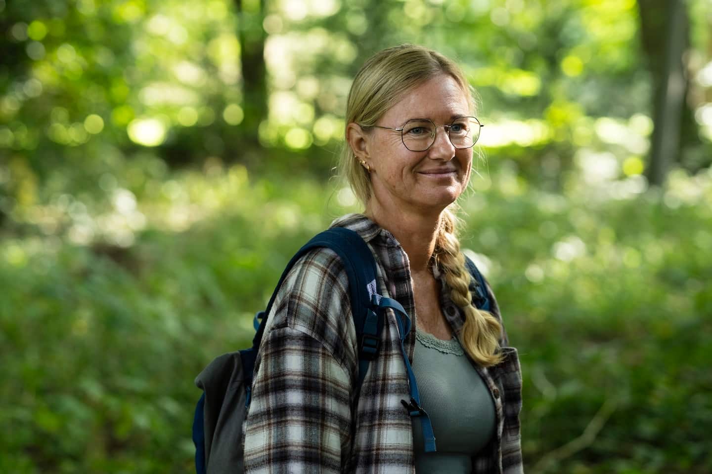 Vrouw met bril en rugzak, lachend in een bosrijke omgeving met groene bladeren op de achtergrond.
