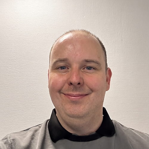 Man met grijze trui en zwarte kraag glimlacht voor een muur met het logo van een bandenbedrijf.