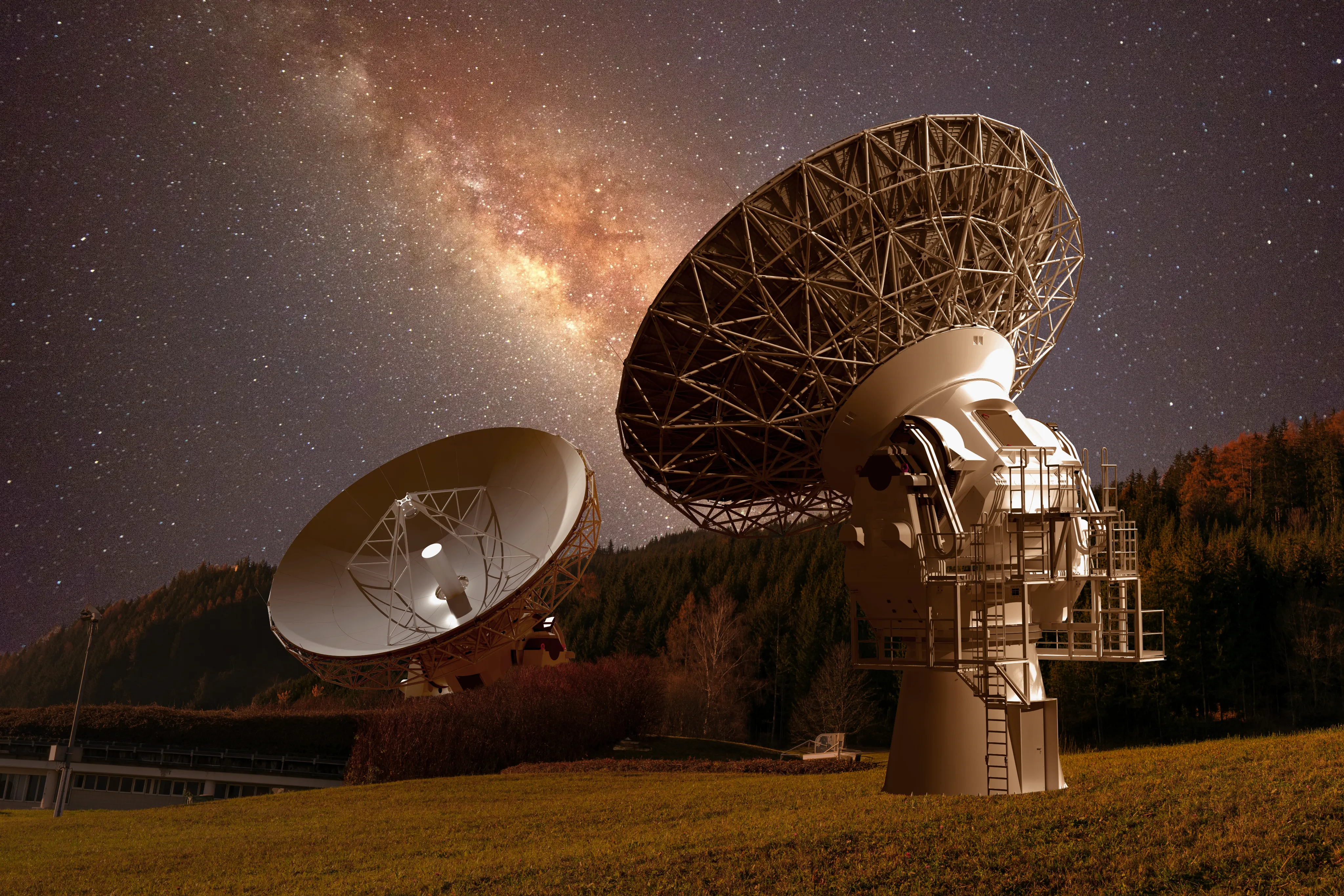 Two large radio telescope dishes in a grassy field with a forest in the background under a starry night sky featuring the Milky Way galaxy.