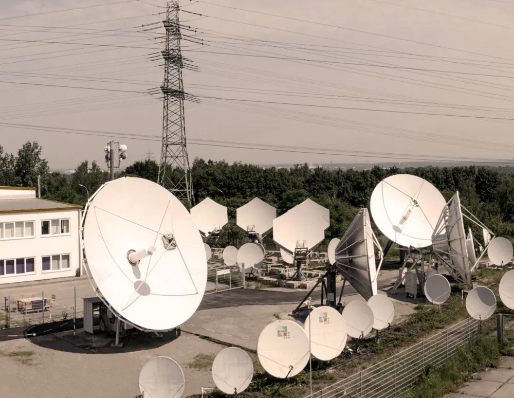 Outdoor satellite communication station with multiple large white satellite dishes and antennas, surrounded by trees and power lines in the background.