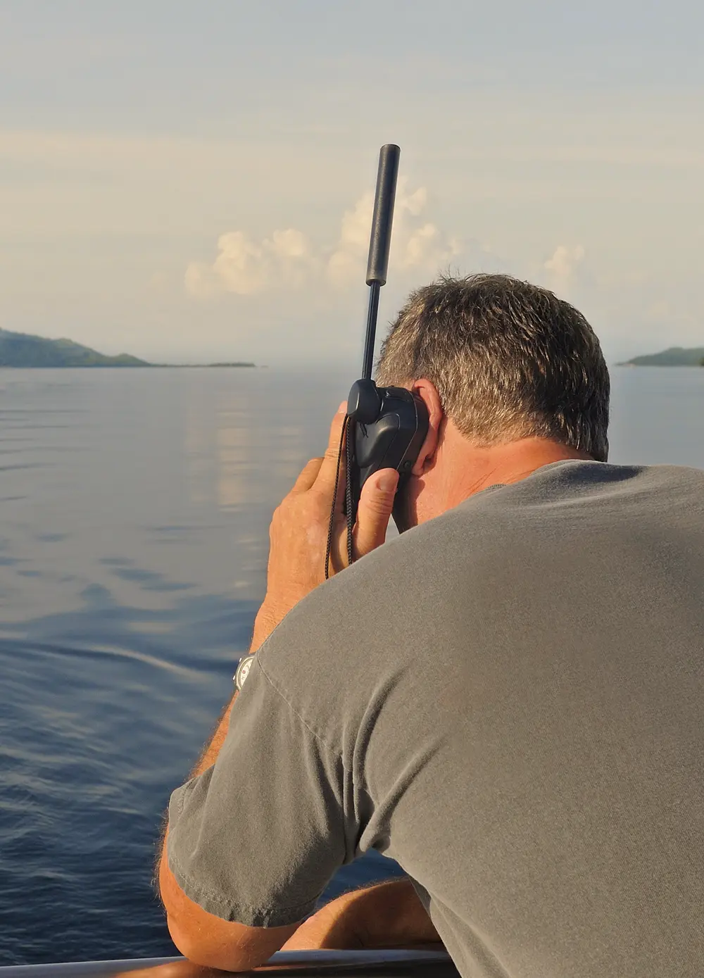 Man with short dark hair wearing a gray T-shirt and watch, holding a black walkie-talkie to his ear while sitting by calm water with distant land and cloudy sky.