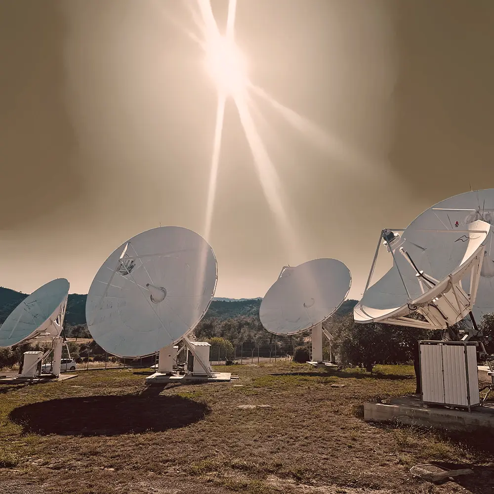 Array of large white satellite dishes on mounts in an open grassy area with hills and trees in the background, under a bright sun with lens flare in the sky.