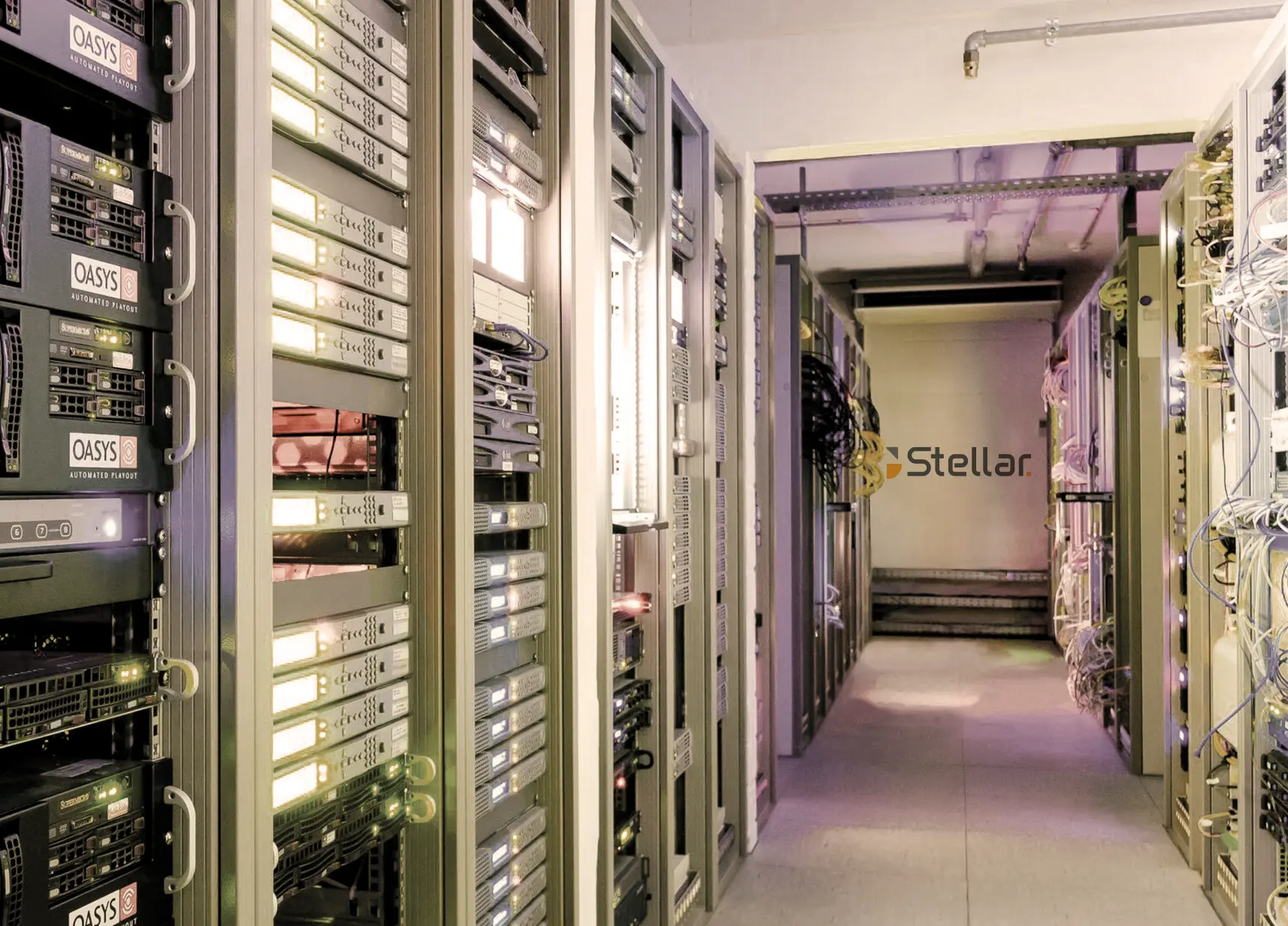 Interior view of a data center corridor with server racks on both sides filled with various network and computing equipment, featuring cables and blinking lights, and a wall at the end with the word 'Stellar' displayed.