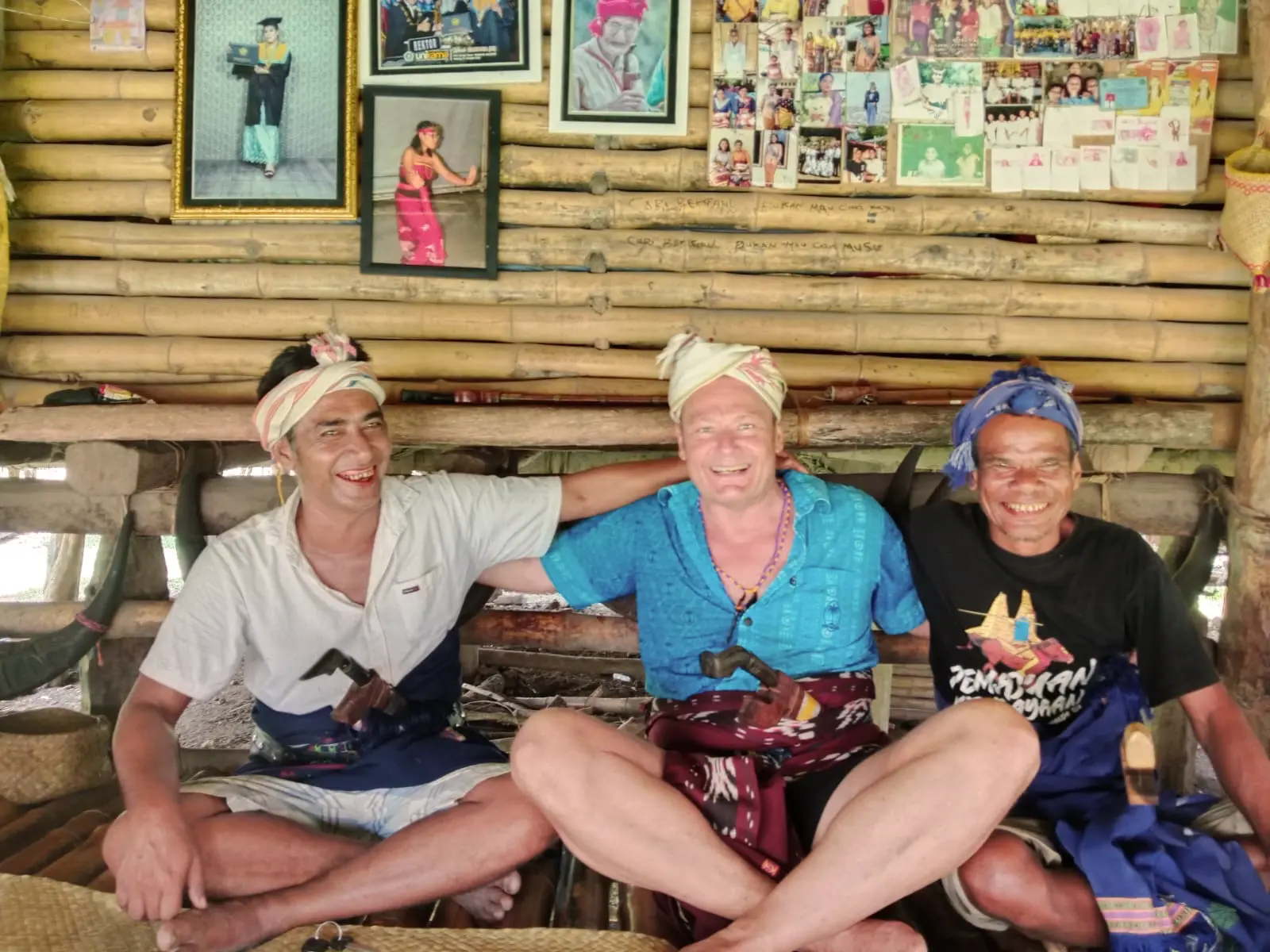 Three men sitting cross-legged on a woven mat inside a bamboo structure, smiling and posing for a photo; the two men on the sides wear headscarves and traditional cloth belts holding curved knives, while the man in the middle wears a blue shirt and a similar belt and headscarf. Behind them are framed pictures and a wall covered with photographs and drawings.