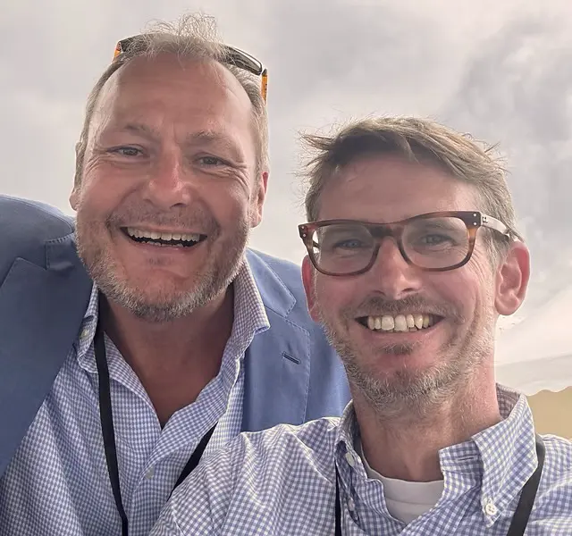 Two men smiling at the camera, one wearing glasses and both dressed in checkered shirts and jackets, with a cloudy sky background.