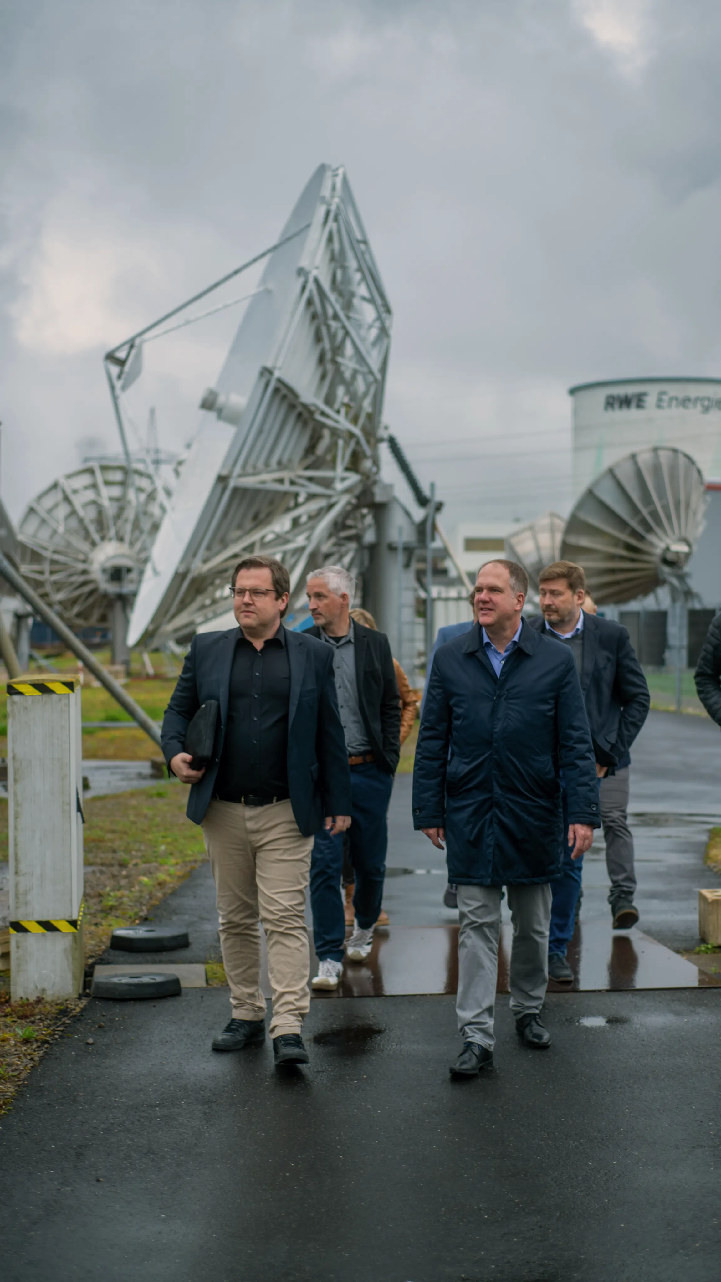 Group of business people walking outside on a wet pavement near large satellite dishes with cloudy sky above.