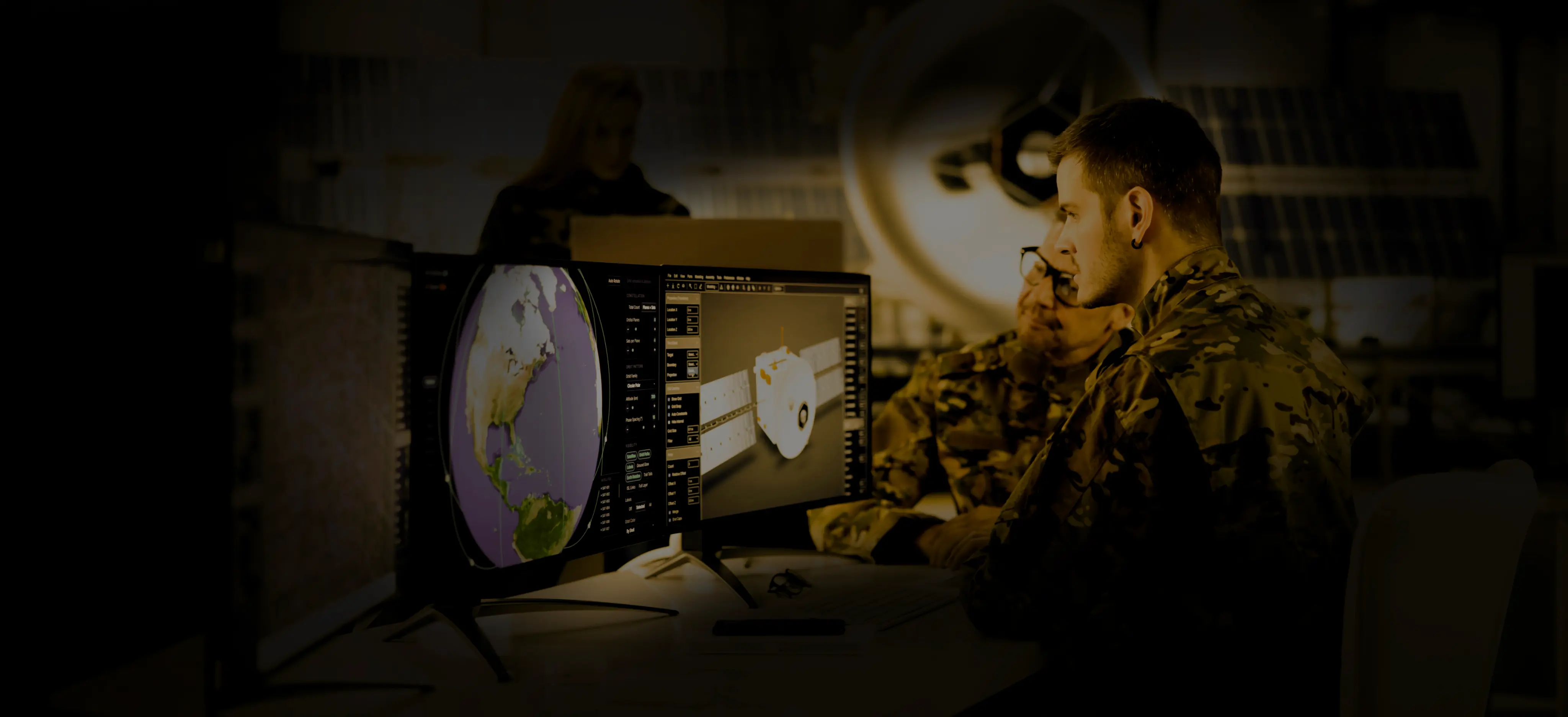 Two military personnel in camouflage uniforms intently analyzing satellite data on dual monitors, one displaying a globe focused on the Americas and the other showing a satellite model, with a blurred figure in the background.