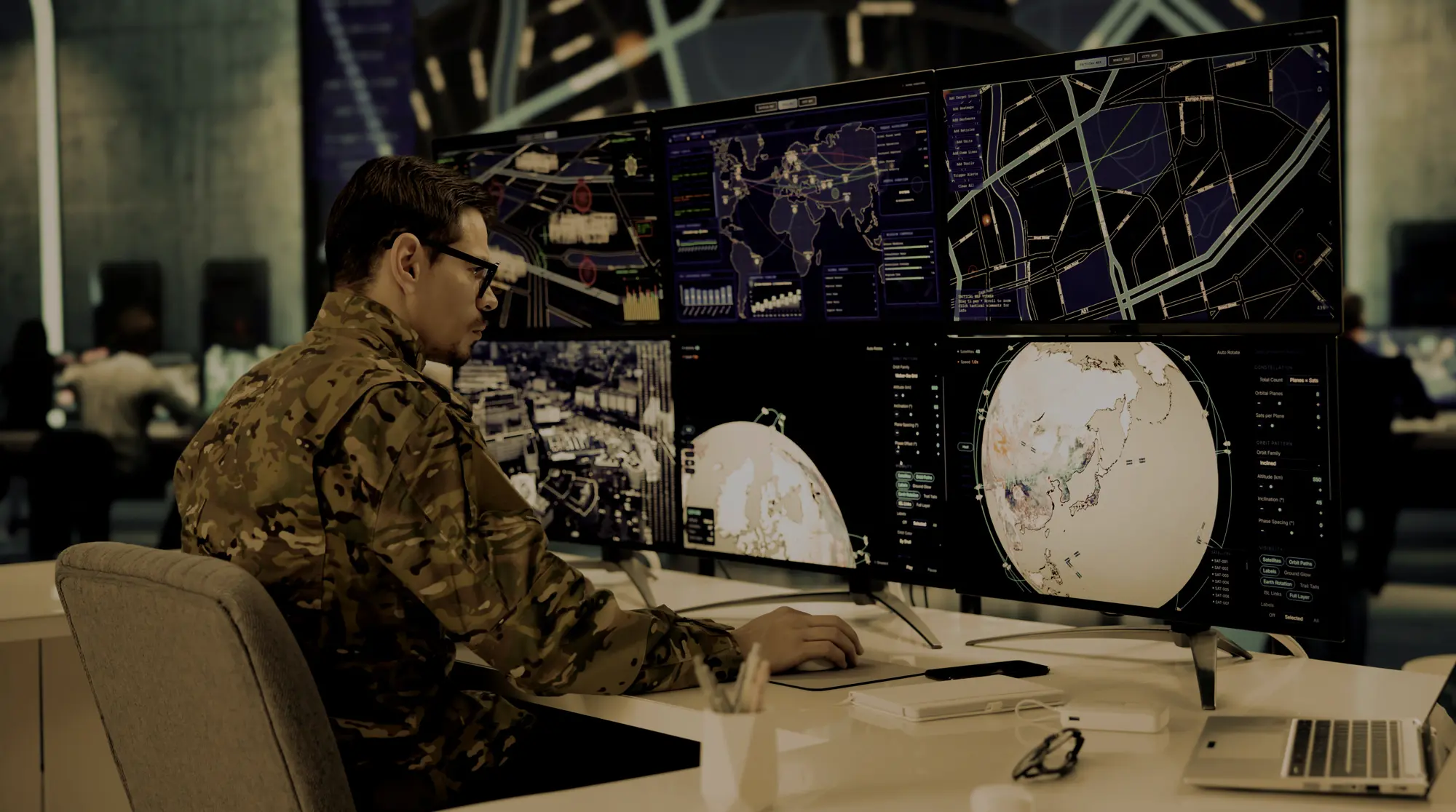 A man in camouflage military uniform and glasses sits at a desk with four large monitors displaying global maps, satellite data, and navigation information, in a control room environment.