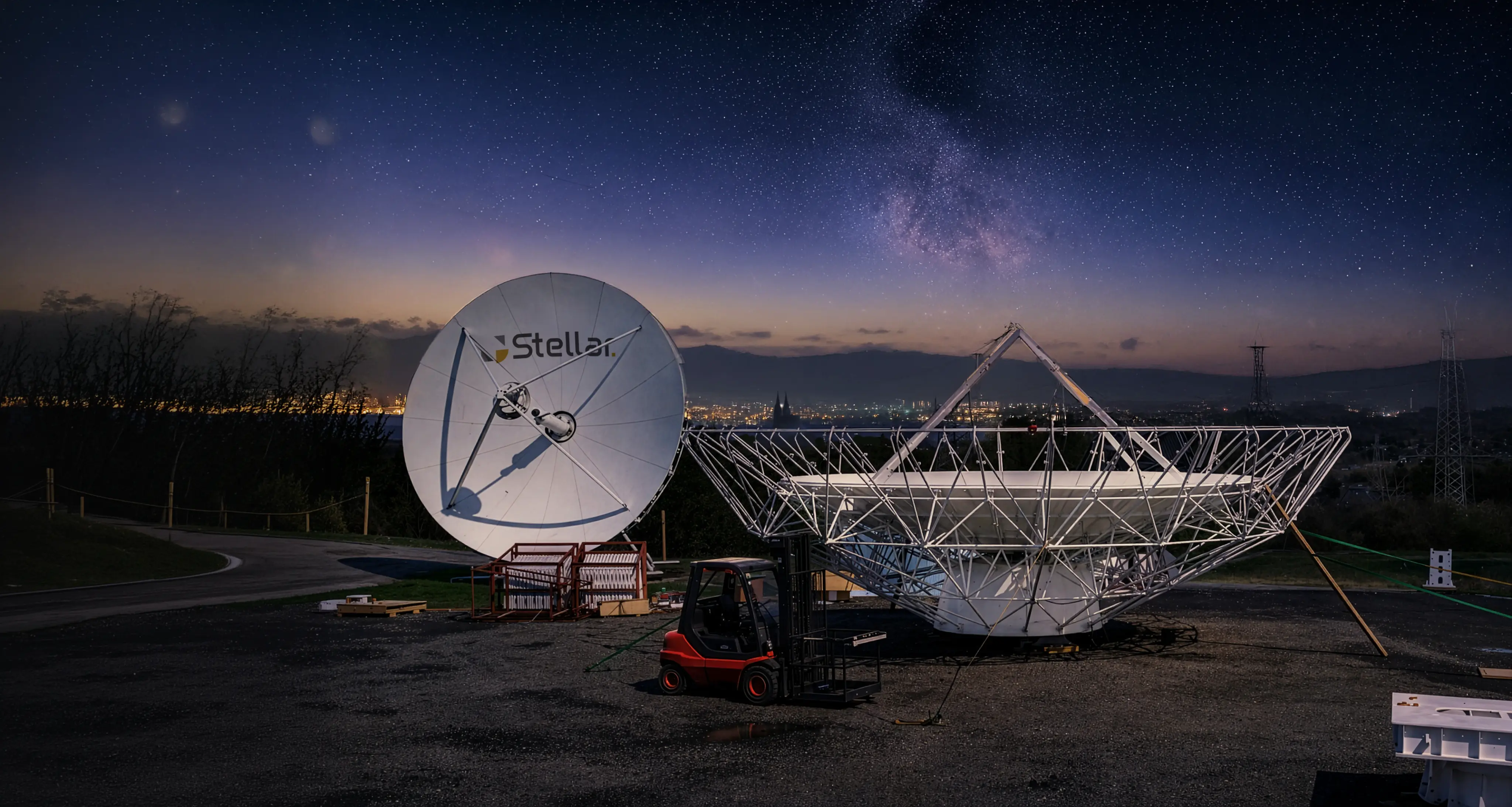 Two large satellite dishes outdoors at dusk with a forklift nearby, city lights and starry sky with the Milky Way in the background.