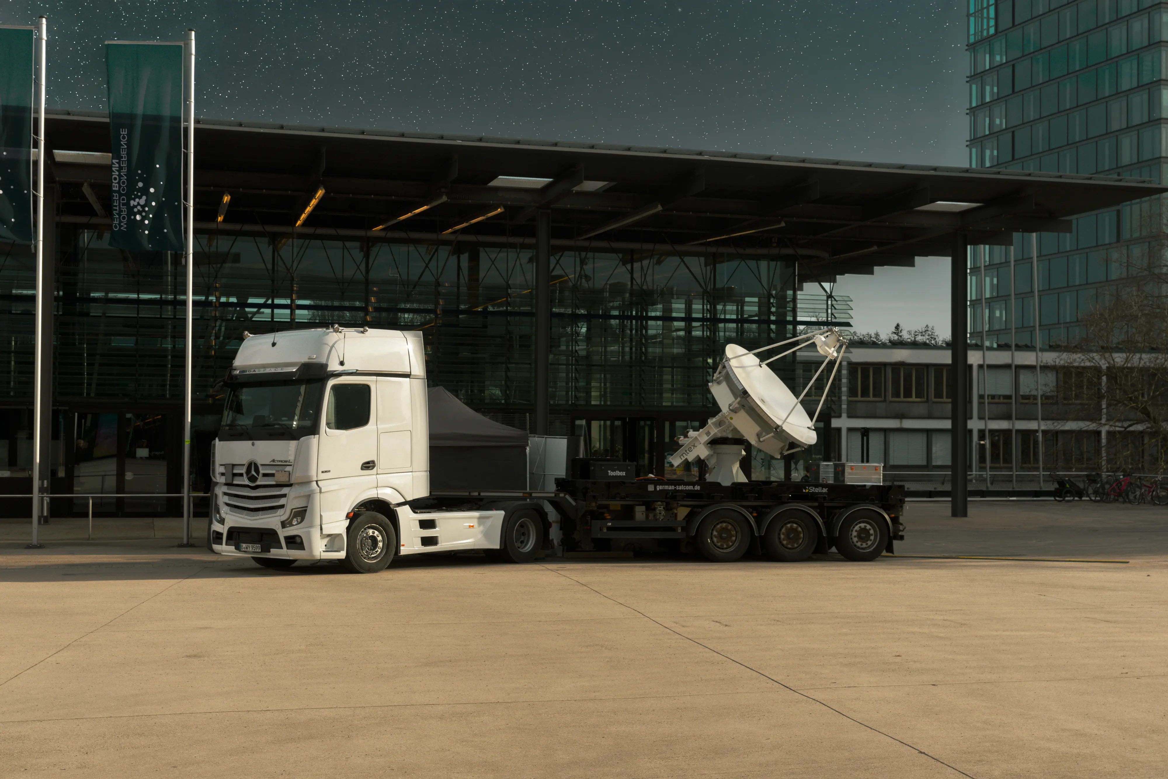 White Mercedes-Benz Actros truck parked in front of a modern glass building, carrying a large satellite dish on its trailer during the evening under a starry sky.