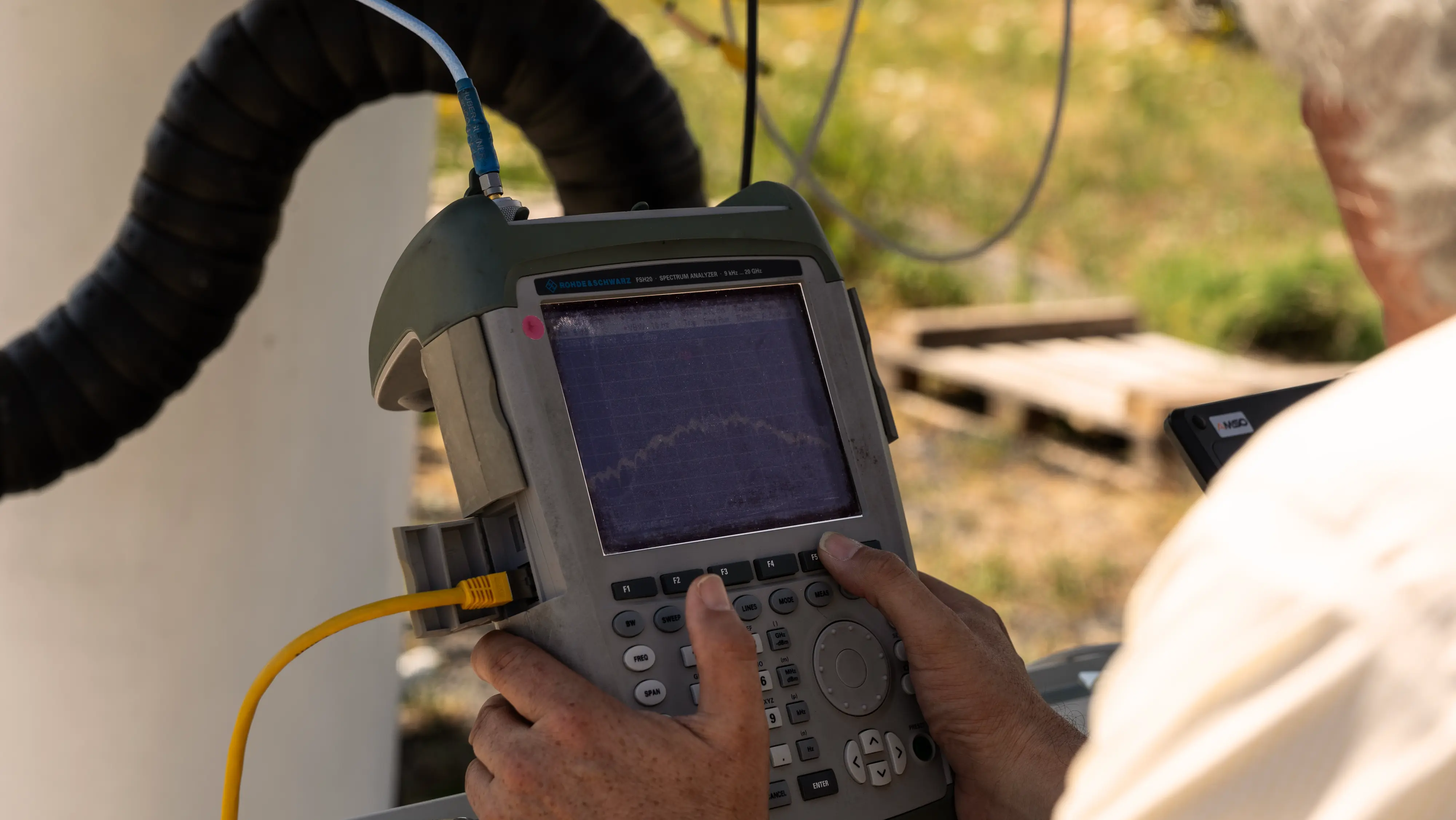 Person operating a Rohde & Schwarz FSH3 spectrum analyzer outdoors, adjusting controls with both hands, with a yellow cable connected to the device.
